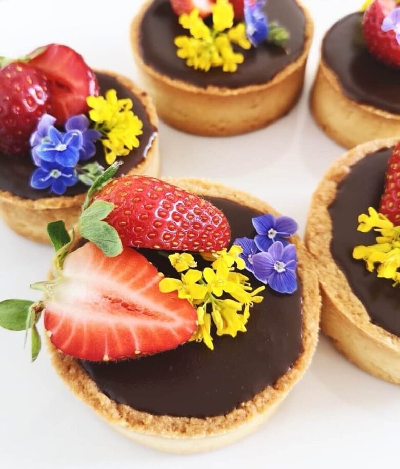Šokolādes mini tartes ar spīdīgu franču ganašu, svaigām zemenēm un pļavas ziediem uz gaiša fona